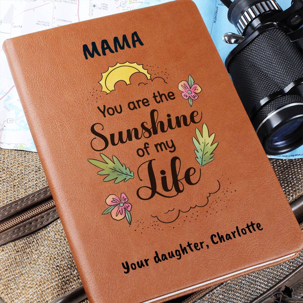 Brown notebook with 'Mama, you are the sunshine of my life' text and floral designs, placed on a textured surface with a camera in the background.
