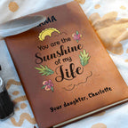 Brown leather notebook with 'You are the Sunshine of my Life' text and floral designs on a patterned fabric background.