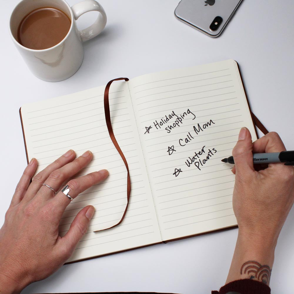 Person writing in a notebook with a cup of coffee and phone on a white surface